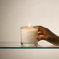 Hand holding a lit candle in a glass container with 'Plantas Medicinas' branding on a neutral background.